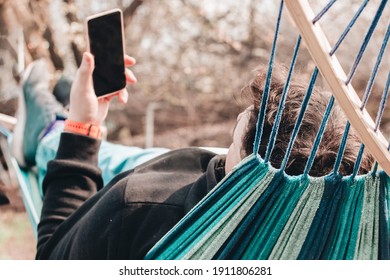 Young Man Lying In Hammock In Back Yard Holding Smartphone In Hand. Male In Blue Jeans And Black Sweater Lies In Hammock In Garden Working, Looking At His Phone.  Work From Home, Relaxing Concept