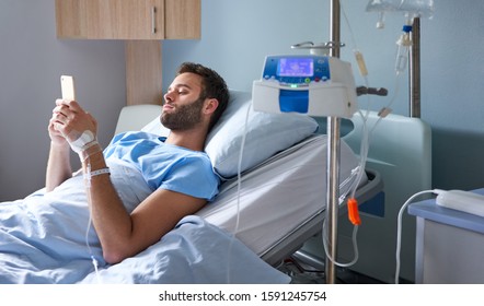 Young man lying in a hospital bed getting an intravenous drip treatment and using his cellphone - Shutterstock ID 1591245754