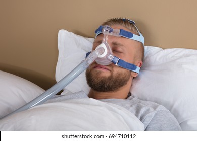 Young Man Lying On Bed With Sleeping Apnea And CPAP Machine