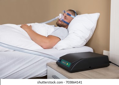 Young Man Lying On Bed With Sleeping Apnea And CPAP Machine