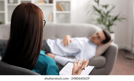 A young man is lying on a couch at a psychologist's appointment. A brunette female psychologist in glasses conducts a psychological session sitting on a chair in her office. - Powered by Shutterstock