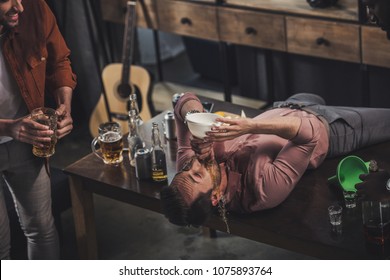 Young Man Lying On Table And Drinking Beer From Funnel While Friends Looking At Him