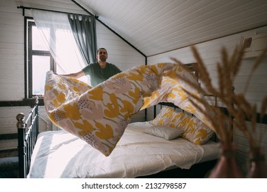 A Young Man Makes A Bed In A Cozy Bedroom At Home. A Handsome Guy In Home Clothes Does House Cleaning, Changes Bed Linen, Straightens A Blanket On A Large Bed In A Bright Room.