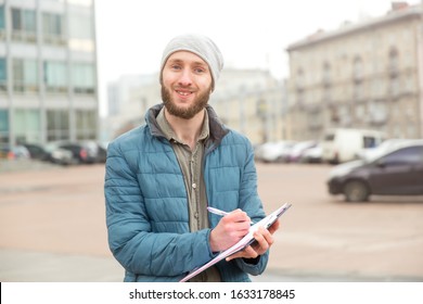 Young Man Makes Records Of Research Or Questioning Against The Background Of A City Street. Public Opinion Poll, Collection Of Statistical Data, Questioning.