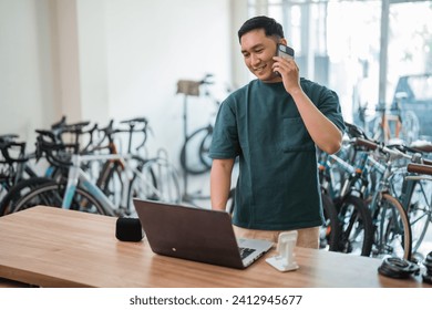 young man making calls while working on a laptop at a desk in a bicycle shop - Powered by Shutterstock