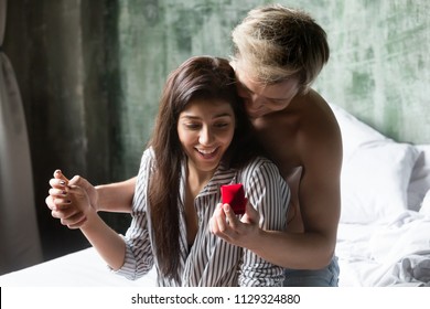 Young Man Making Marriage Proposal To Excited Beautiful Girlfriend Sitting On Bed In Morning, Happy Millennial Woman Getting Unexpected Engagement Ring Present When Boyfriend Propose To Her In Bedroom