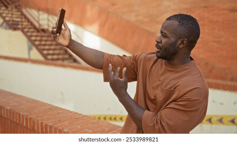 Young man making a video call in an urban setting, holding a smartphone outdoors against a brick wall on a sunny day, appearing engaged and expressive in conversation. - Powered by Shutterstock