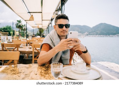 Young Man Male In Sunglasses Looking At His Mobile Smart Phone Gadget In A Street Sea Side Cafe Restaurant Coffee Shop With Scenery Mountains In The Background. Hello Summer Holiday Vacation.