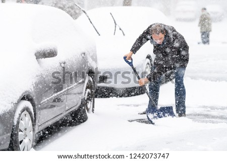 Young man, male in winter coat cleaning, shoveling driveway, street from covered falling, snow in heavy snowing snowstorm with shovel, residential houses, cars parked on road