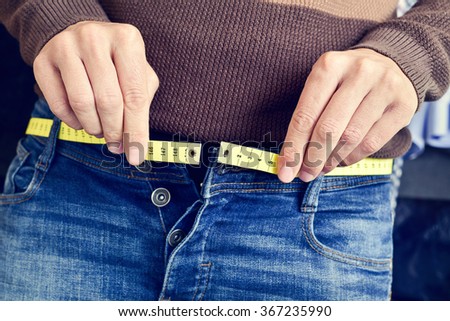 a young man with a measuring tape as a belt tries to fasten his trousers, because of the weight gain