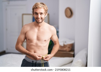 Young Man Measuring Waist Using Tape Measure At Bedroom