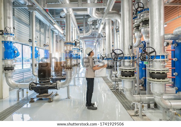 Young man mechanical engineer holding drawing to\
checking and inspection of HVAC heating ventilation air\
conditioning system and pipping line of industrial construction at\
boiler pump room system