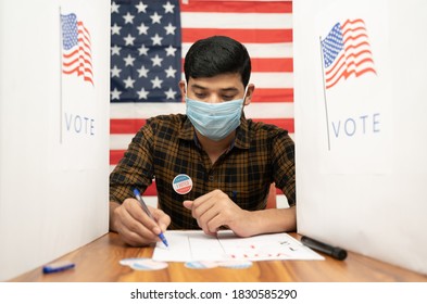 Young Man In Medical Mask Busy Inside The Polling Booth With US Flag As Background - Concept Of In Person Voting With Covid-19 Safety Measure At US Election