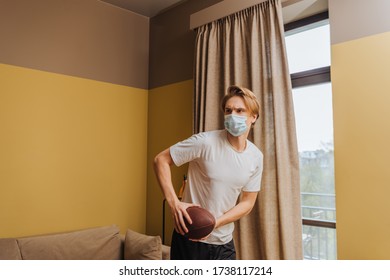 Young Man In Medical Mask Holding American Football In Living Room
