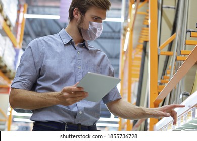 Young Man In Medical Mask Shopping Or Working In A Hardware Warehouse Standing Checking Supplies On His Tablet.