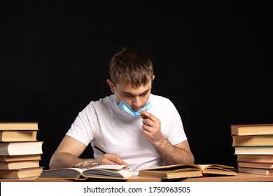 Young Man In A Medical Mask Sits At A Table With Documents And Books. Student Is Preparing For The Exam. Quarantined Distance Learning.