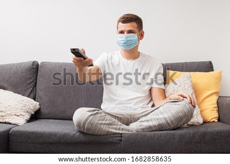 Young man in medical mask sits on couch and wathing television during quarantine. Coronavirus, stay home