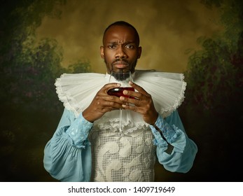 Young Man As A Medieval Grandee Or Nobleman On Dark Studio Background. Holding A Glass Of Red Wine. Portrait In Retro Costume. Human Emotions, Comparison Of Eras And Facial Expressions Concept.