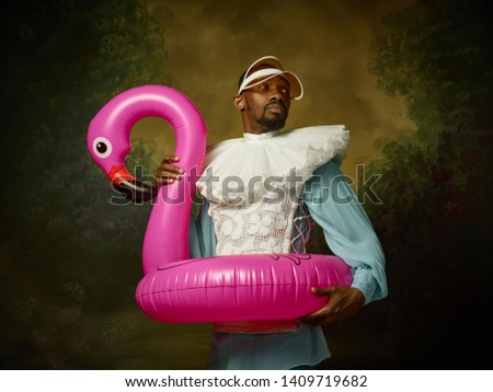 Young man as a medieval grandee or nobleman in a swimring as a pink flamingo on dark studio background. Portrait in retro costume. Human emotions, comparison of eras and facial expressions concept.