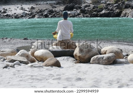 A young man meditates on the banks of the Ganges River in Rishikesh, India. Photo from behind.