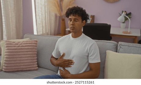 Young man meditating with hands on stomach sitting on a couch in a well-lit living room with neutral tones and modern decor. - Powered by Shutterstock