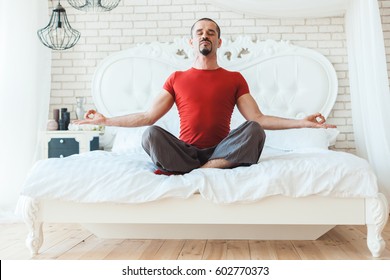 Young Man Meditating In The Morning Sitting On The Bed In The Room.