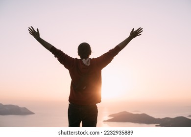 A young man meets the sunset, arms outstretched in a free gesture on a mountain by the sea. A Caucasian guy stands high on a cliff above a small town and looks at the sun setting behind the islands - Shutterstock ID 2232935889