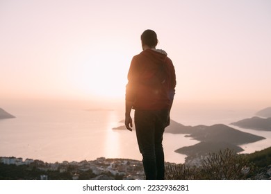 A young man meets a sunset on a mountain by the sea. A Caucasian guy stands high on a cliff above a small town and looks at the sun setting behind the islands - Shutterstock ID 2232936281