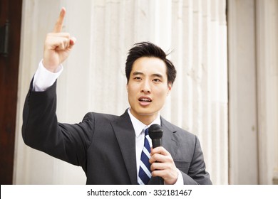 A Young Man With A Microphone And His Finger In The Air Giving A Speech.