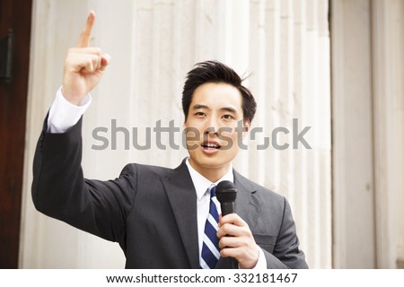 A young man with a microphone and his finger in the air giving a speech.