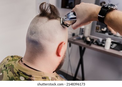 A young man in a military uniform shaves his head bald for military service. A guy with a beard gets a haircut at a barber shop. - Shutterstock ID 2148396173