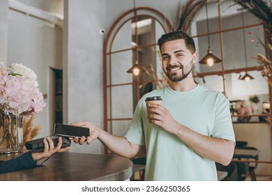 Young man in mint t-shirt buy takeaway paper cup coffee bar counter hold wireless modern bank payment terminal use mobile phone nfc indoors cafe Urban leisure lifestyle catering establishment concept - Powered by Shutterstock