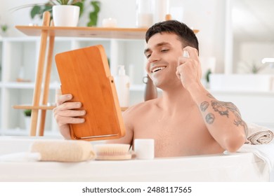 Young man with mirror applying facial product in bathtub at home - Powered by Shutterstock