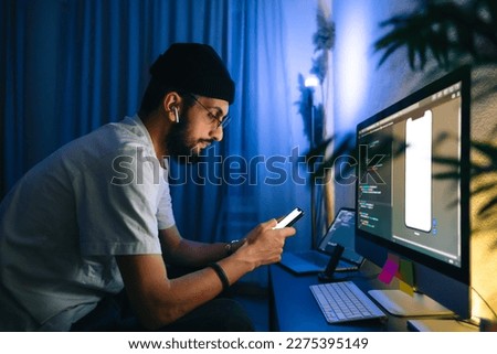 Young man mobile developer writes program code on a computer and check app in smartphone, programmer work in home office.