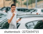 young man with mobile phone looking at cars with expression of success