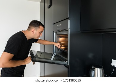 Young Man In Modern Kitchen Leaning Towards Open Oven Door Holding A Fork Looking Past Camera.