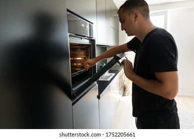 Young Man In Modern Kitchen Leaning Towards Open Oven Door Holding A Fork Looking Past Camera.