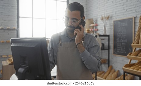 Young man with moustache in bakery shop talking on phone while wearing an apron and standing behind the counter with bread displayed and light streaming through a large window. - Powered by Shutterstock