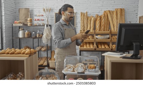 Young man with moustache wearing an apron using a mobile phone in a bakery filled with various breads and pastries on display. - Powered by Shutterstock