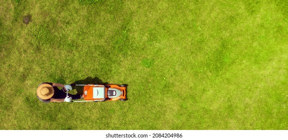 A young man is mowing a lawn with a lawn mower in his beautiful green floral summer garden. A professional gardener with a lawnmower cares for the grass, view from above - Shutterstock ID 2084204986