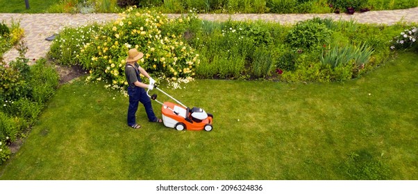 A young man is mowing a lawn with a lawn mower in his beautiful green floral summer garden. A professional gardener with a lawnmower cares for the grass, view from above - Shutterstock ID 2096324836