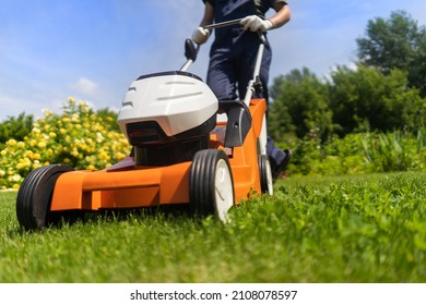 A young man is mowing a lawn with a lawn mower in his beautiful green floral summer garden. A professional gardener with a lawnmower cares for the grass in the backyard. - Shutterstock ID 2108078597