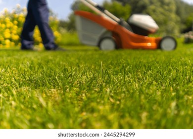 A young man is mowing a lawn with a lawn mower in his beautiful green floral summer garden. A professional gardener with a lawnmower cares for the grass in the backyard, meadow closeup. - Powered by Shutterstock