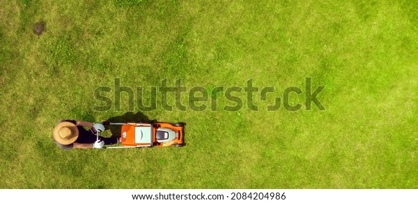 A young man is\
mowing a lawn with a lawn mower in his beautiful green floral\
summer garden. A professional gardener with a lawnmower cares for\
the grass, view from above
