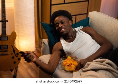 Young Man Muscular Athletic Relaxes In Evening On Couch Watching TV And Eating Chips From Bowl, Student Switches Channels, Programs With Remote Control