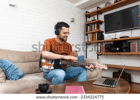 Young man music teacher giving guitar lessons to students online on laptop computer from home. Learning to play electric guitar using online course and listening melody through wireless headphones.
