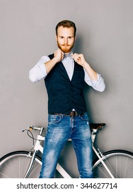 A young man with mustache and beard is near fashionable modern fixgear bicycle. Jeans and shirt, vest and straightens the bow tie hipster style. Gray background. Toned color. Looking at the camera