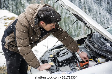 Young Man Near Car With Open Hood Inspecting Engine In Winter. Snowy Forest On Background