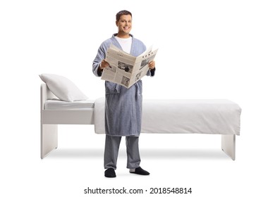 Young Man With A Newspaper Wearing A Bathrobe And Standing Next To A Bed Isolated On White Background