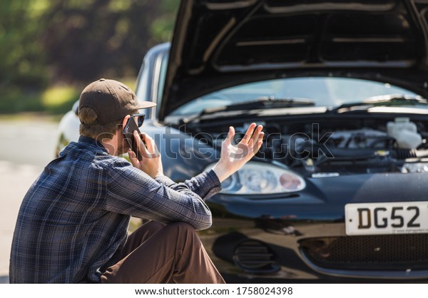 Young man next to his car, depressed and angry on the\
mobile phone telephone call after the car has broken down on the\
roadside waiting for assistance to arrive and recover or fix the\
vehicle 